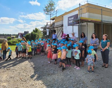 Zahlreiche Kindergartenkinder mit blauen Bauer-Holzbau-Kappen feiern gemeinsam mit ihren Erzieherinnen und weiteren Gästen das Richtfest des Kindergartenanbaus in Gröningen. Im Hintergrund ist der eingerüstete Holzbau mit einem Banner von Bauer Holzbau zu sehen.