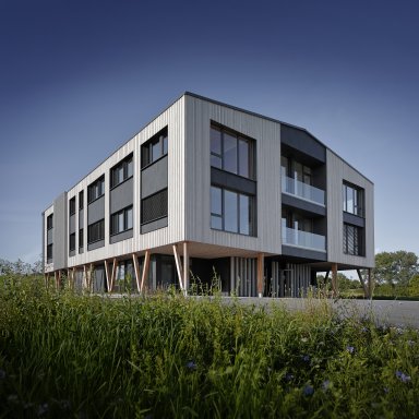 Modernes Verwaltungsgebäude von Bauer Holzbau in Satteldorf-Gröningen mit vertikaler Holzfassade, großen Fensterflächen und dunklen Fassadenelementen. Das Obergeschoss wird von schräg angeordneten Holzstützen getragen. Zwei verglaste Balkone und eine begrünte Außenanlage ergänzen die Architektur.