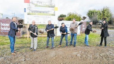 Sieben Projektbeteiligte, darunter Bürgermeister Tobias Strauß, Vertreter des Landkreises, der Kirchengemeinde und des Bauprojekts, stehen beim symbolischen Spatenstich für die neue Kindertagesstätte in Schnelldorf mit Schaufeln auf dem Baugrundstück. Im Hintergrund sind die Bautafel und ein Banner von Bauer Holzbau zu sehen.