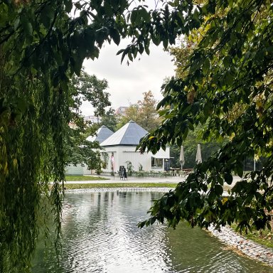 Blick durch die Bäume über den Teich auf den neuen Kiosk KÖSK im Unteren Schlossgarten Stuttgart. Die markanten Pyramidendächer des Gebäudes ragen zwischen den Bäumen hervor.
