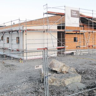 Rohbau der neuen Kindertagesstätte Schwalbennest in Schnelldorf während der Bauphase. Das eingeschossige Gebäude in Holzbauweise ist eingerüstet und teilweise mit Fassadenplatten verkleidet. Am Baugerüst sind Banner von Bauer Holzbau angebracht.