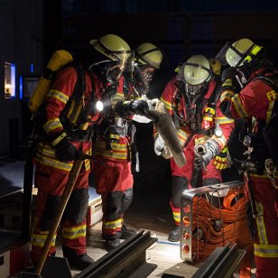 Mehrere Feuerwehrleute mit Atemschutz arbeiten während der gemeinsamen Übung in einer Fertigungshalle von Bauer Holzbau mit Schläuchen und Einsatzgeräten.